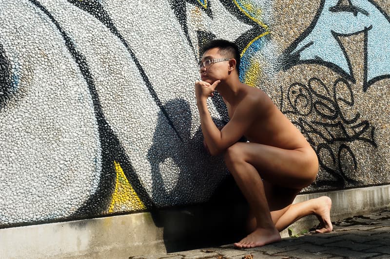 A nude young man with glasses sits thoughtfully on the sidewalk against a vibrant graffiti wall, one hand on his chin, lost in contemplation as sunlight casts a long shadow behind him.